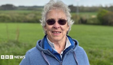 Kate has curly grey hair and round sunglasses. She wears a blue hooded top and stands in front of the fields where the solar farm will go.