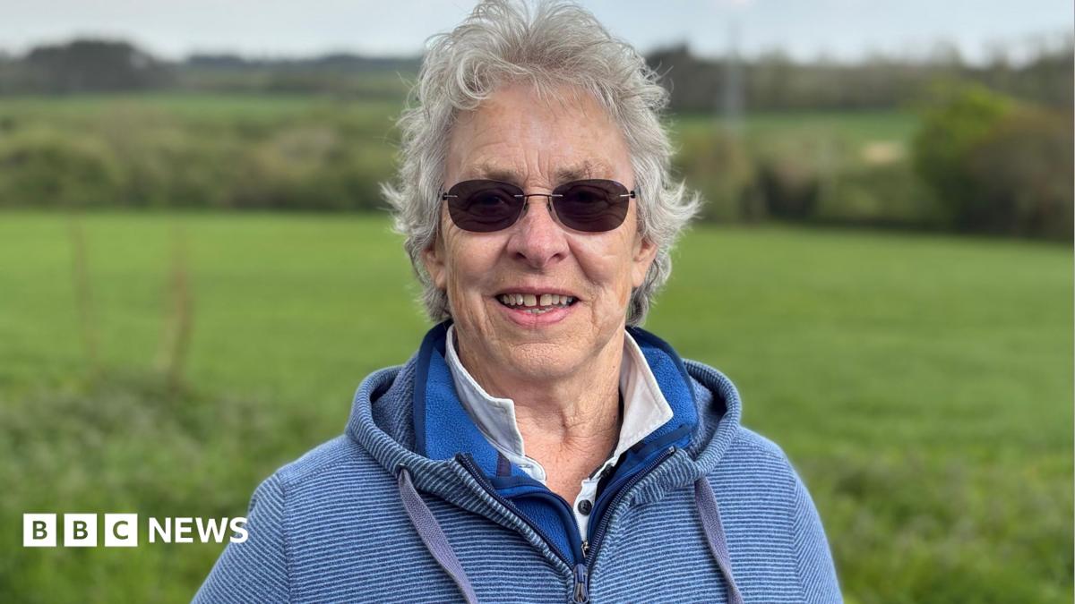 Kate has curly grey hair and round sunglasses. She wears a blue hooded top and stands in front of the fields where the solar farm will go.