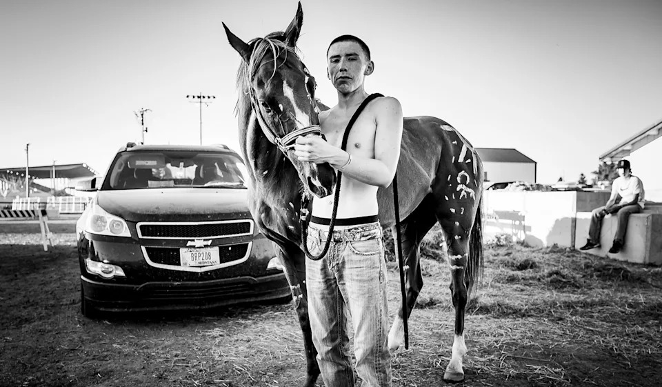 a man and his horse pose together in a hay-covered parking lot
