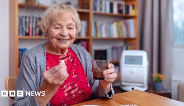 An elderly woman wearing a red floral dress and grey cardigan. She is holding a glass of water and a pill. She is sat at a wooden table. Behind her is a white wall and bookshelf.