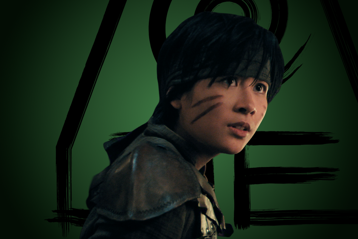 Wes Valarao as Smellerbee with short dark hair, face paint, and leather armor stands in front of a dark green background with bold black shapes, suggesting a fantasy or adventure setting.