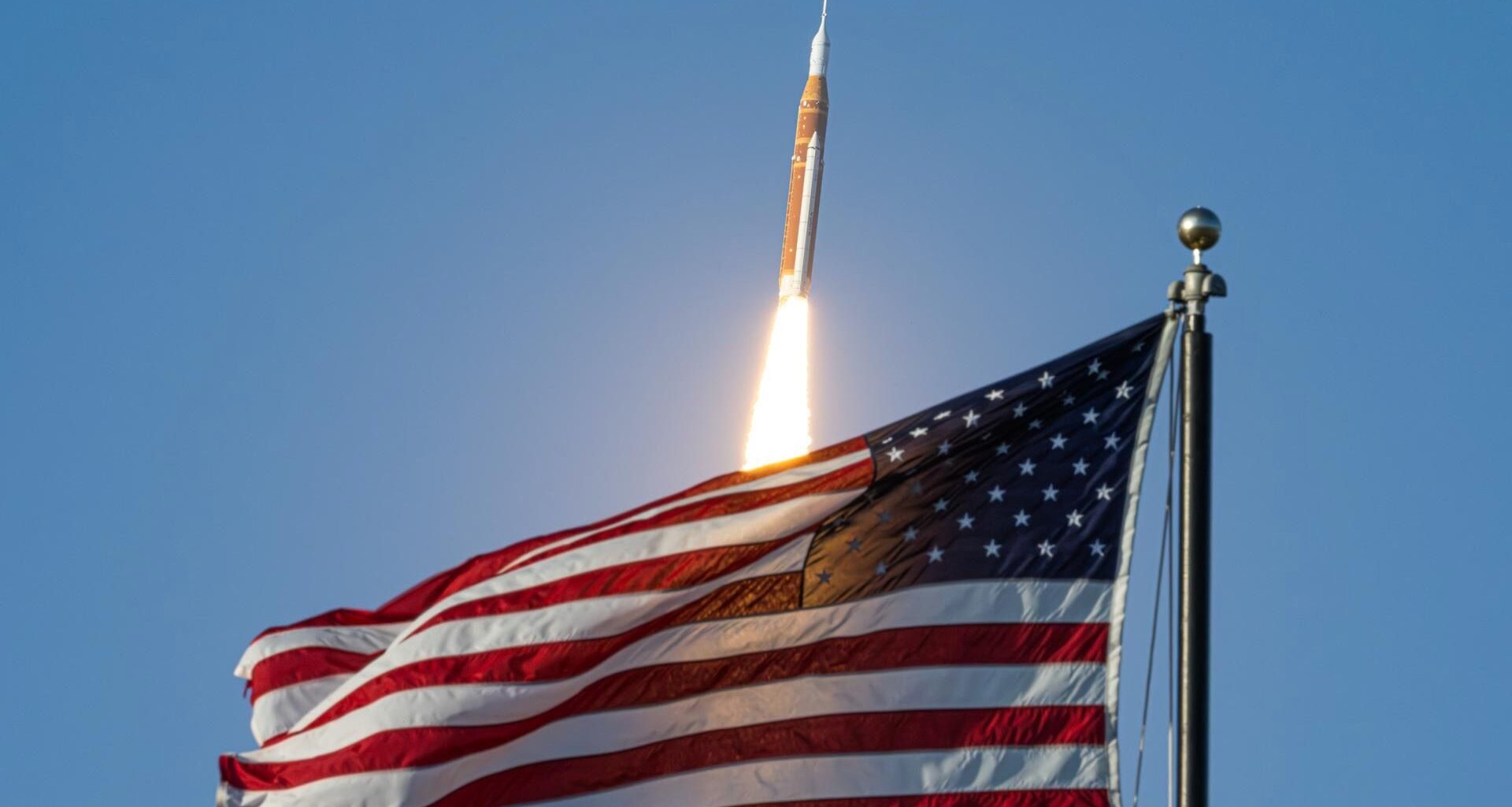 a big orange and white rocket launches into a blue sky with a big american flag in the foreground