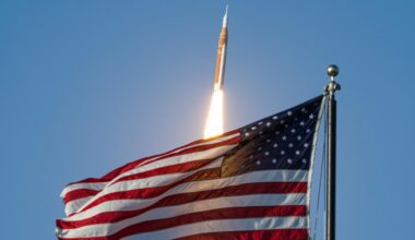 a big orange and white rocket launches into a blue sky with a big american flag in the foreground