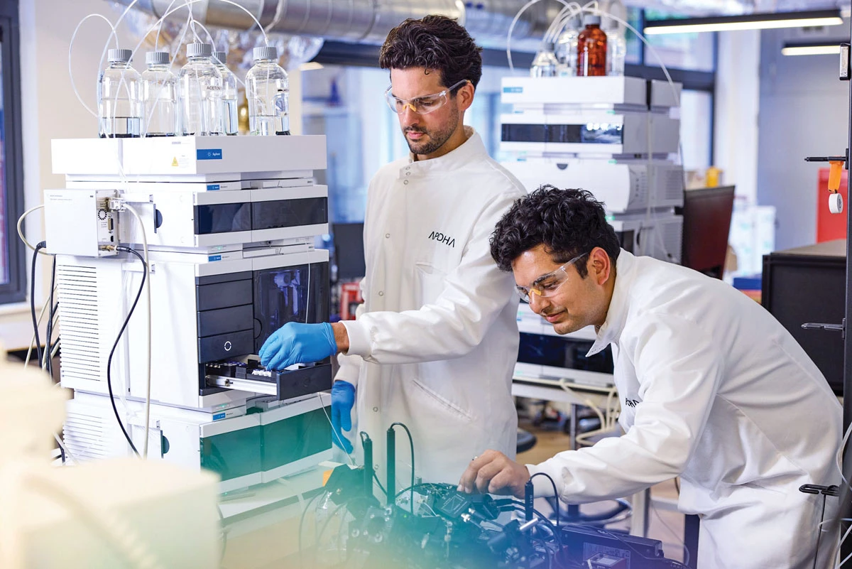 Two scientists in lab coats and protective glasses work at a large white machine.