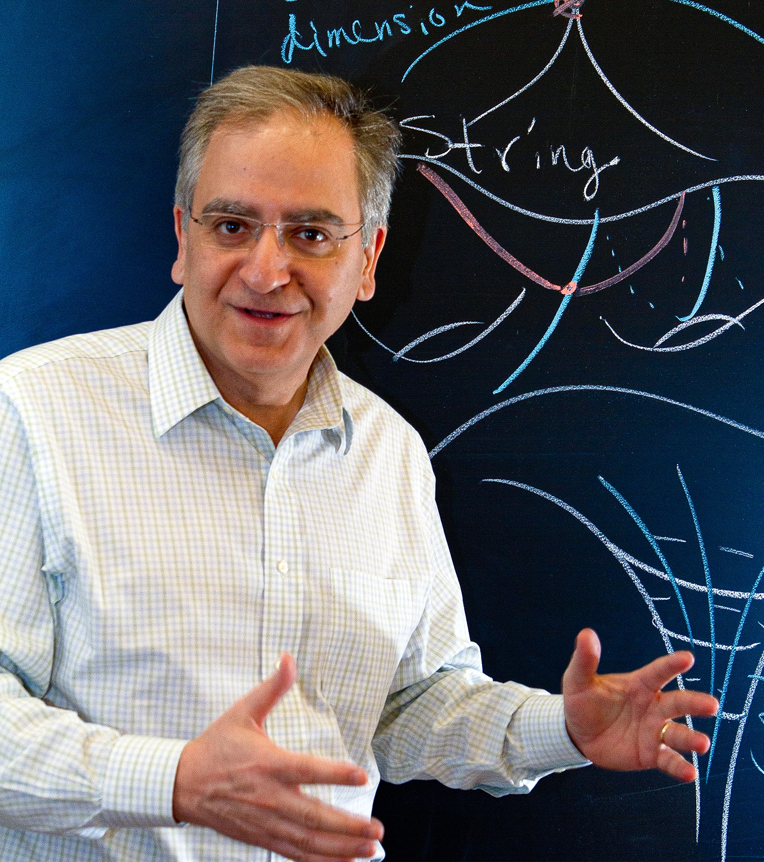 A man stands in front of a blackboard with diagrams and the word “String” written on it.