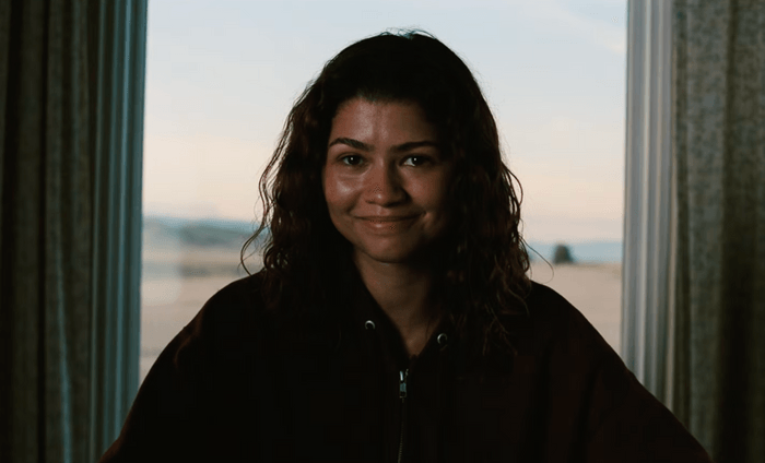 Zendaya smiles while seated in a hoodie as Rue Bennett in Euphoria season 3 episode 8.
