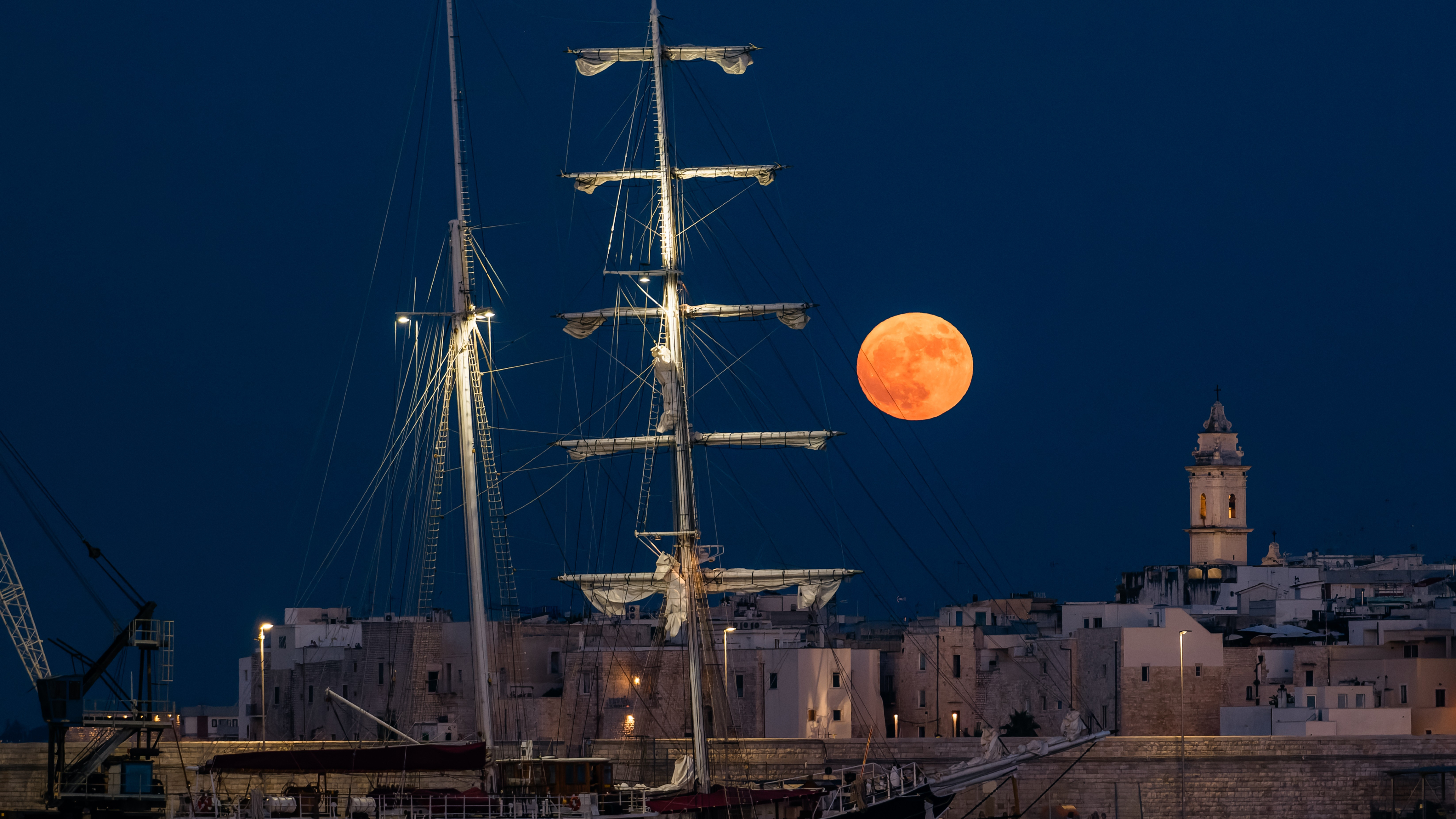 a large full moon with a pinkish orange hue sits low in the sky behind a large sailing boat.