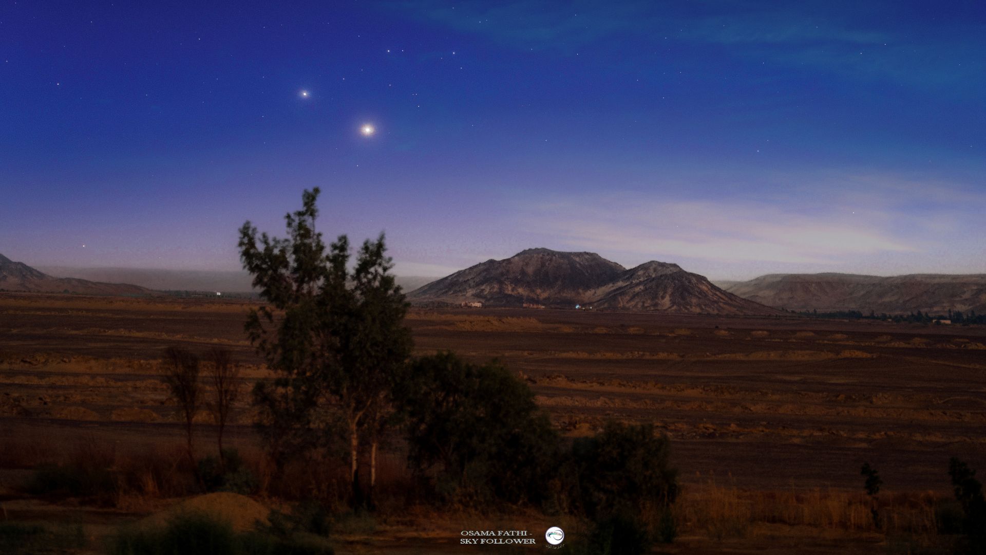 Two bright star-like planets are photographed shining side by side above mountains in a tranquil plain.