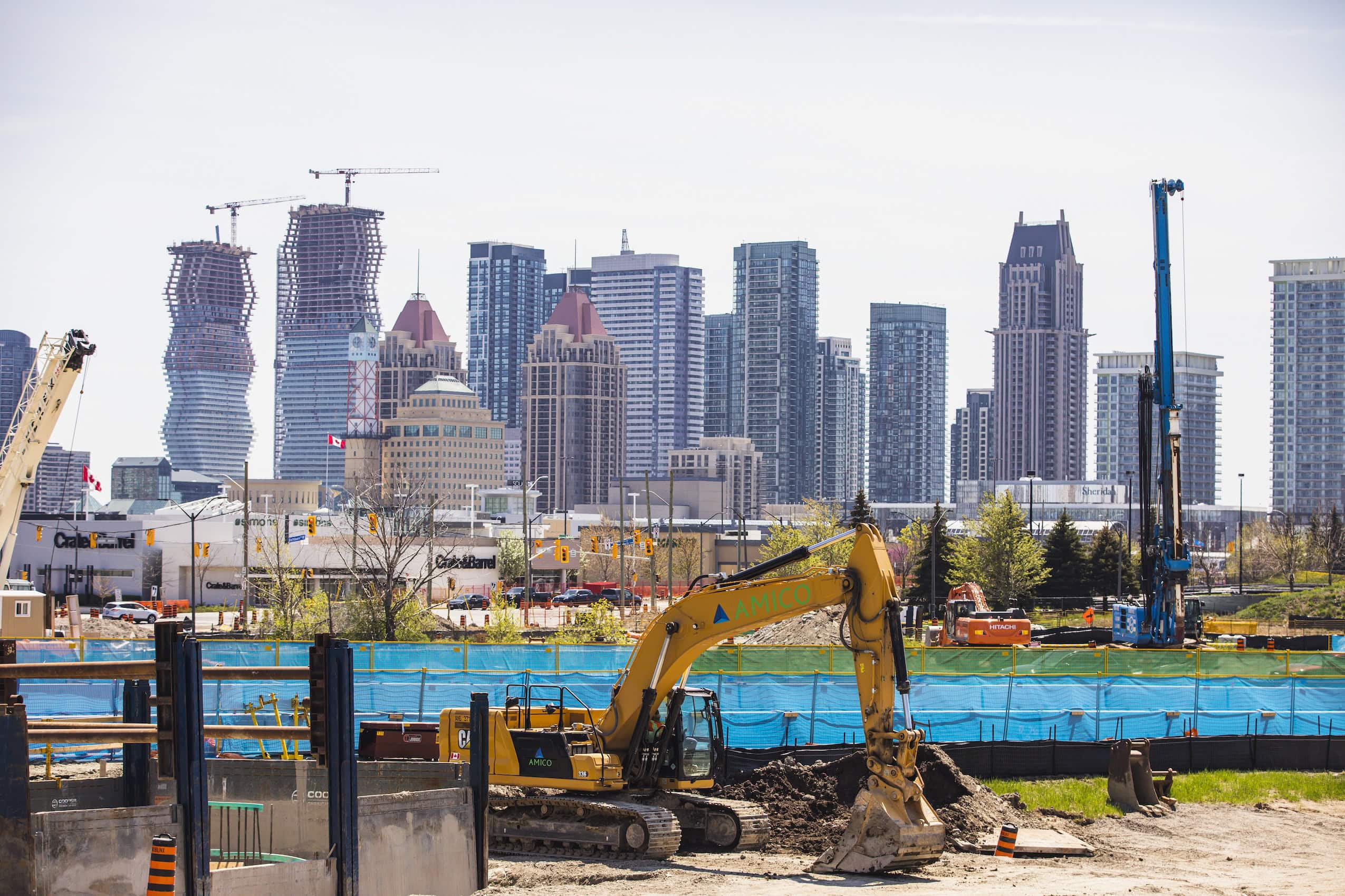 A backhoe at a construction site with a row of skyscrapers, some of them under construction, in the background.