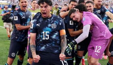Paraguay players celebrate together on the field, fists clenched and shouting, surrounded by teammates and a packed stadium crowd.