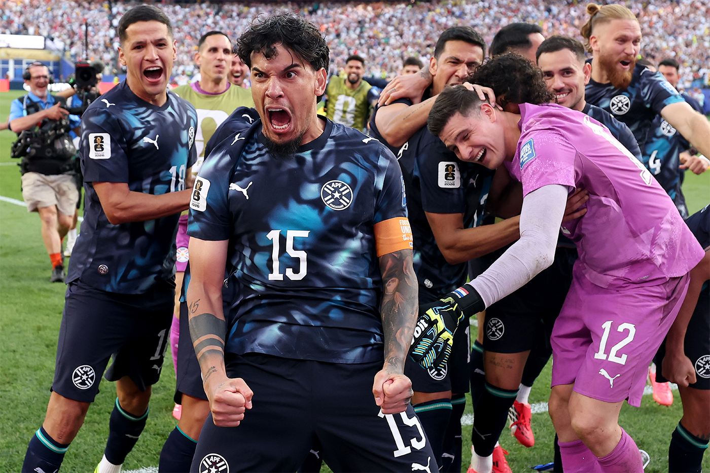 Paraguay players celebrate together on the field, fists clenched and shouting, surrounded by teammates and a packed stadium crowd.