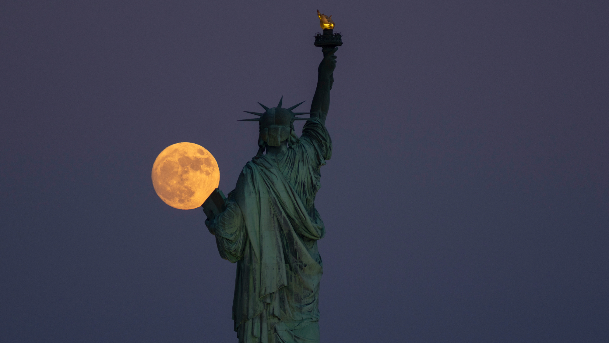 a large fully illuminated moon next to the statue of liberty, it looks like she is holding it in her hand.