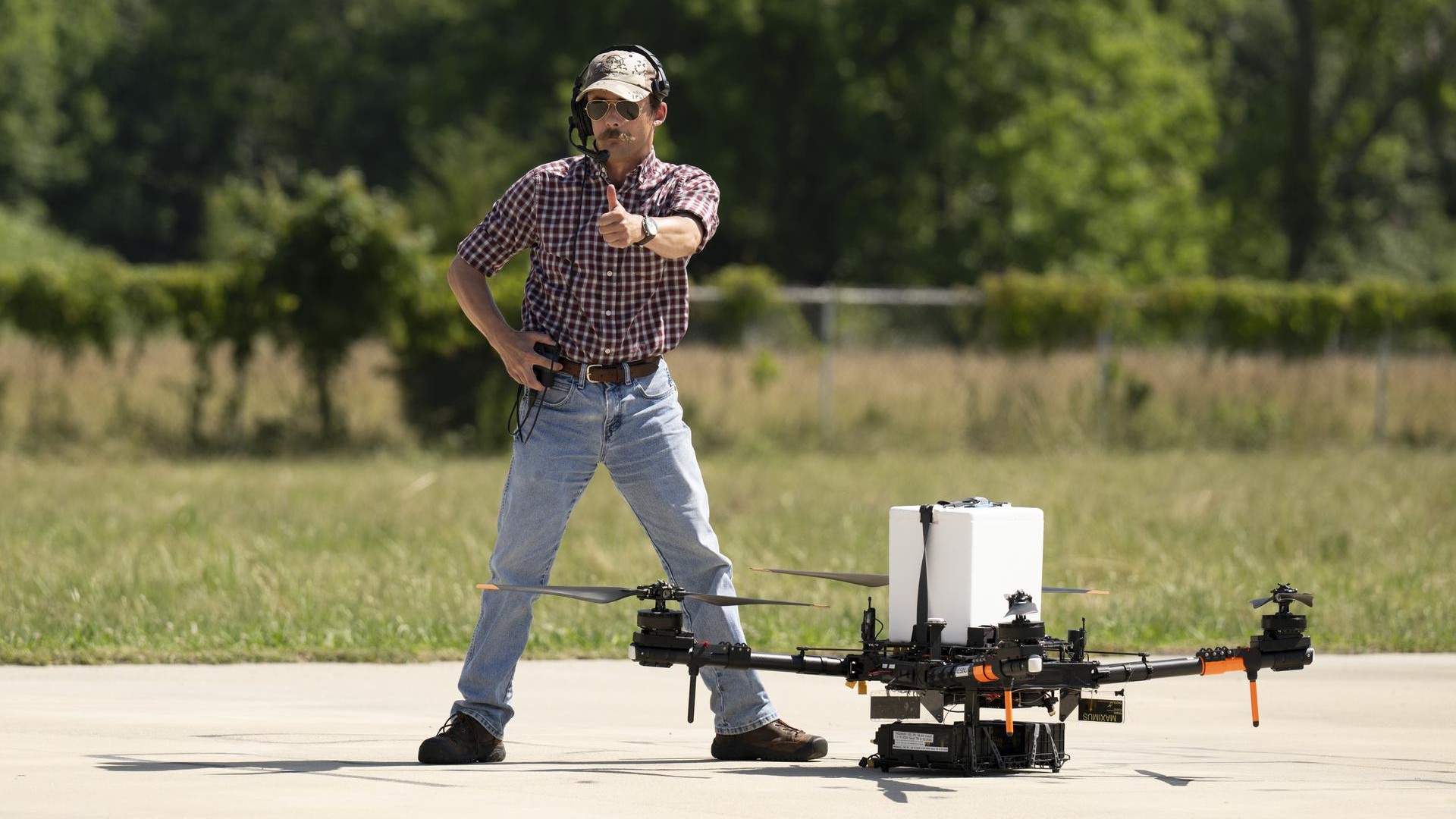 a man with a formidable mustache gives a thumbs up next to a large quadcopter drone resting on a tarmac, with a rectangular white cooler strapped to it