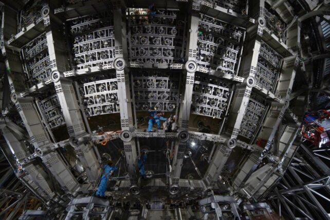 Workers in blue protective suits install components inside the large spherical JUNO detector, surrounded by metal frames and circular sensor mounts.