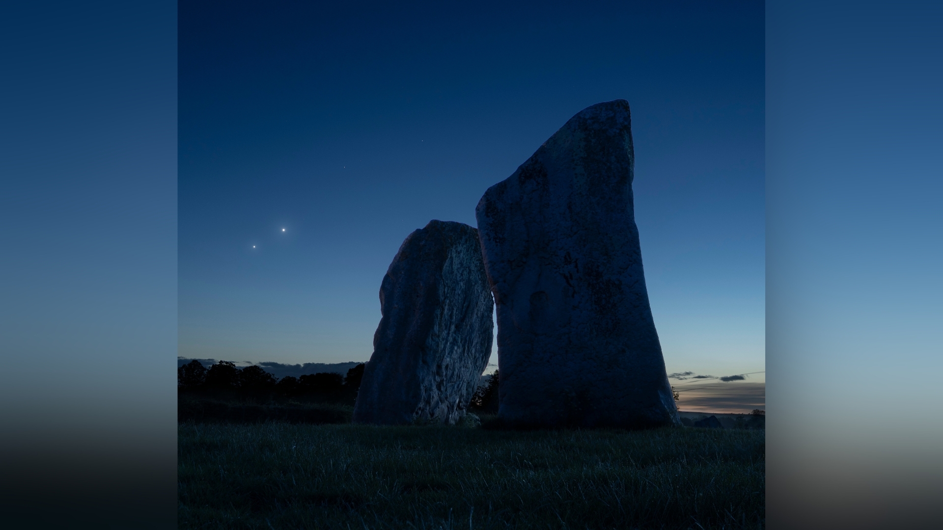 Bright star-like planets are photographed glowing in a dark sky over a pair of large stones, as fainter stars dot the night sky nearby.
