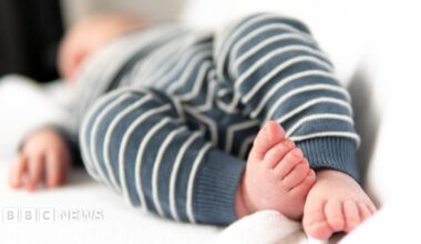 A newborn baby is lying in white sheets, its face is out of focus in the distance, its feet are close to the camera.