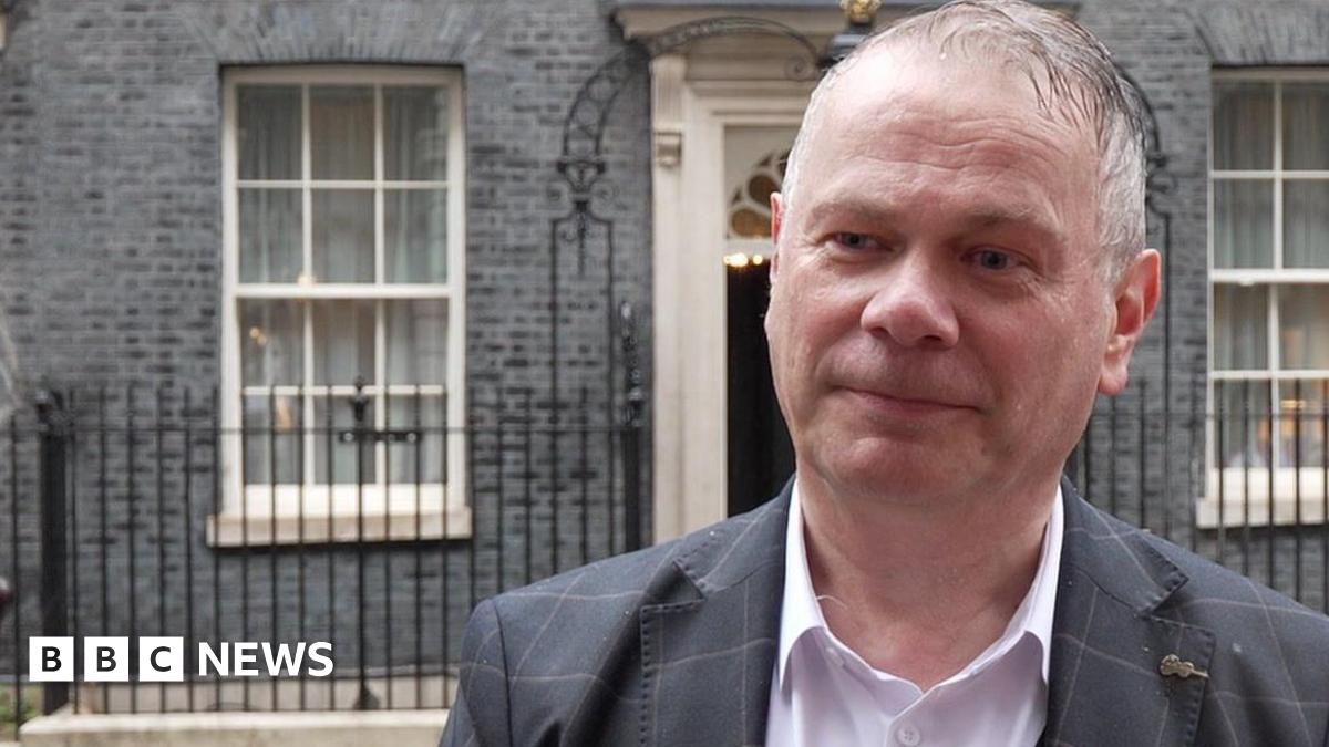 Corin Dalby, with receding grey hair and wearing a white shirt and grey suit with a lighter grey pattern, stands in the rain across the road from 10 Downing Street.