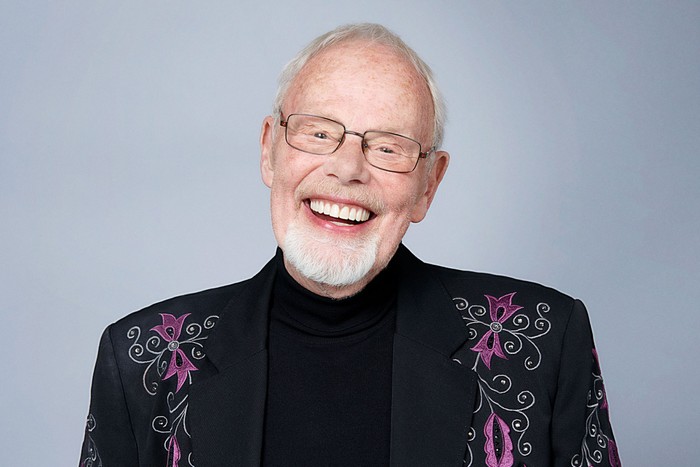 bob-harris Bob Harris wearing a black suit with purple floral designs, smiling ahead in front of a grey background.