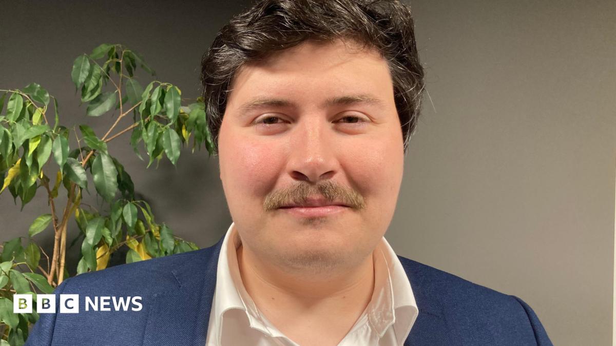 A close up of Deputy Tom Rylatt. He has short black hair, a brown moustache and is wearing a white shirt and navy jacket.