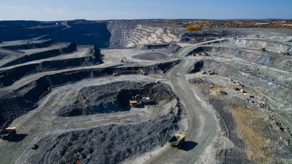 An aerial, wide shot of a large, multi-level open-pit mine. The mine features numerous gray and dark gray rock terraces and winding dirt roads where several yellow excavators and large dump trucks are visible. In the background, a steep, light-colored rock face forms one side of the pit, and beyond it, a distant town with buildings and trees showing autumn foliage can be seen under a bright blue sky.