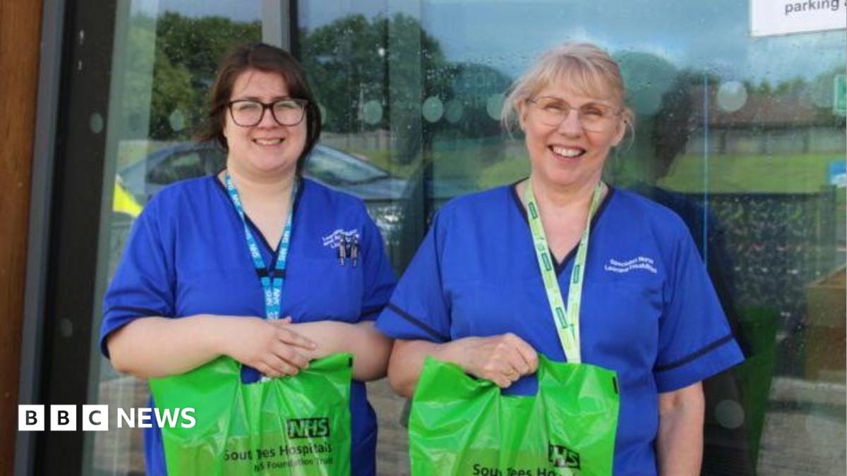 Nurses Elizabeth Dixon and Gemma Crooks standing outside the hospital. They are wearing blue uniforms and holding green care bags. Both wear glasses. One has short brown hair and the other blond hair with a fringe which may be short or tied back.