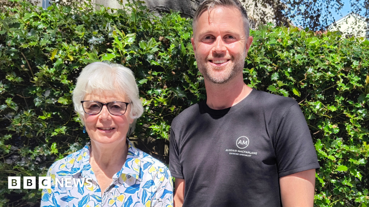 Alastair and his mum Anne are pictured standing in front of a green bush. They are both smiling at the camera. Anne has short white hair and is wearing a white shirt with a blue leaf and yellow flower pattern. She is also wearing black-rimmed glasses. Alastair has short greying hair and stubble. He is wearing a black T-shirt with a white logo.