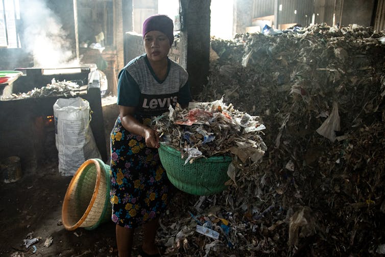 A woman carries a basket with plastic waste from a large pile in a room.