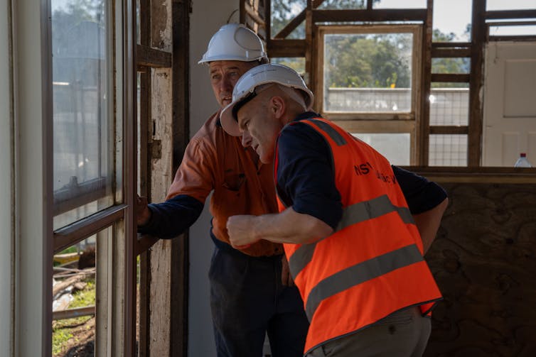Two men in workwear and hard hats study the timber inside a house being deconstructed.