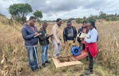 A group of people stands around a hole in the ground with notepads