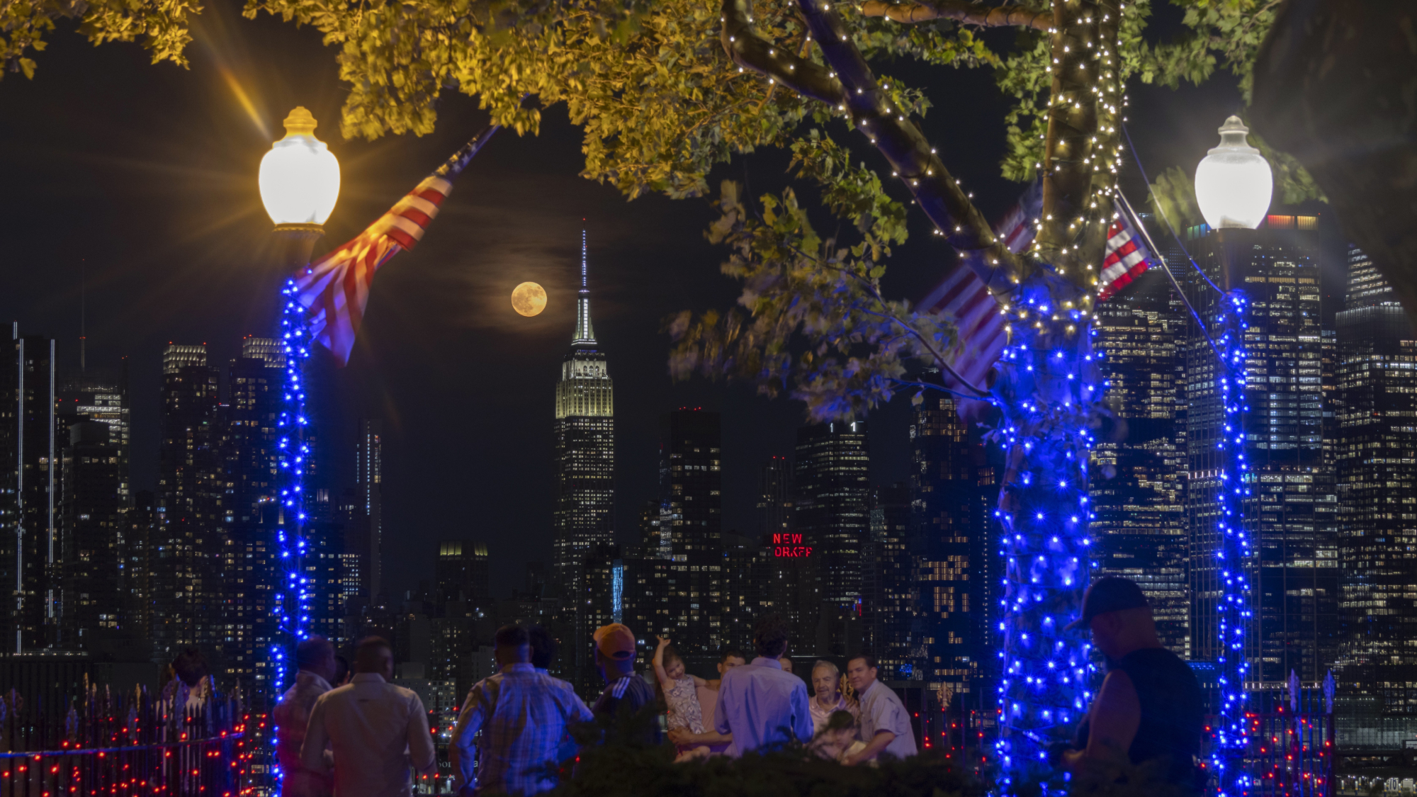 The full moon appears to the left of the Empire State Building, in the foreground, people are sitting outside. There are lampposts adorned with lights and two American flags are visible.