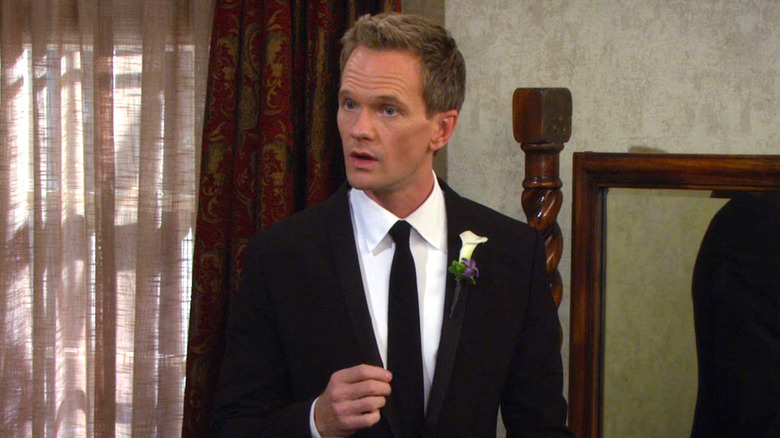 Neil Patrick Harris as Barney Stinson wearing a dark suit, white shirt, and a black tie with a flower pinned to his lapel.