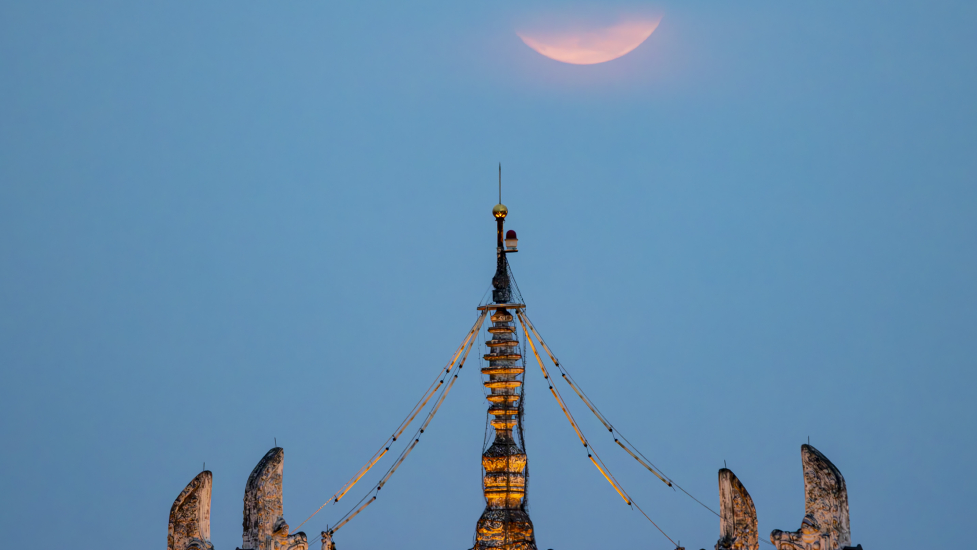 a thin sliver of full moon appearing hazy pink above an ornate rooftop structure.
