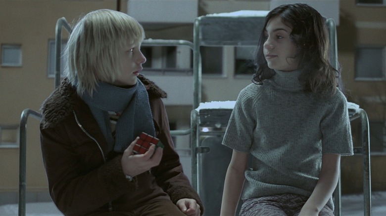 Kåre Hedebrant as Oskar and Lina Leandersson as Eli in Let The Right One In, sitting next to each other outside in the snow against the background of a drab apartment block while Oskar holds a Rubik's Cube..