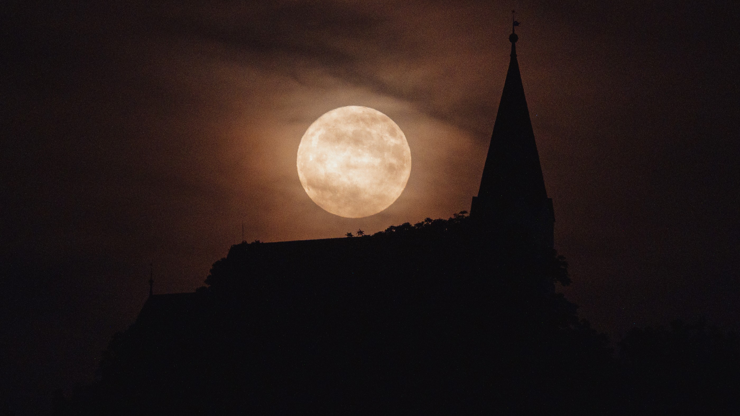 fully illuminated moon behind wispy clouds and the silhouette of a church below.