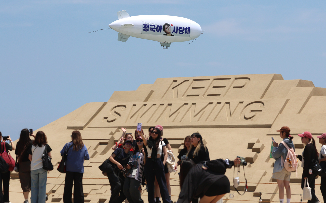 Fans take photos as a blimp carrying a support message for BTS member Jungkook flies above Haeundae Beach in Busan on Friday, behind a sand sculpture inspired by the group’s new song “Swim.” (Yonhap)
