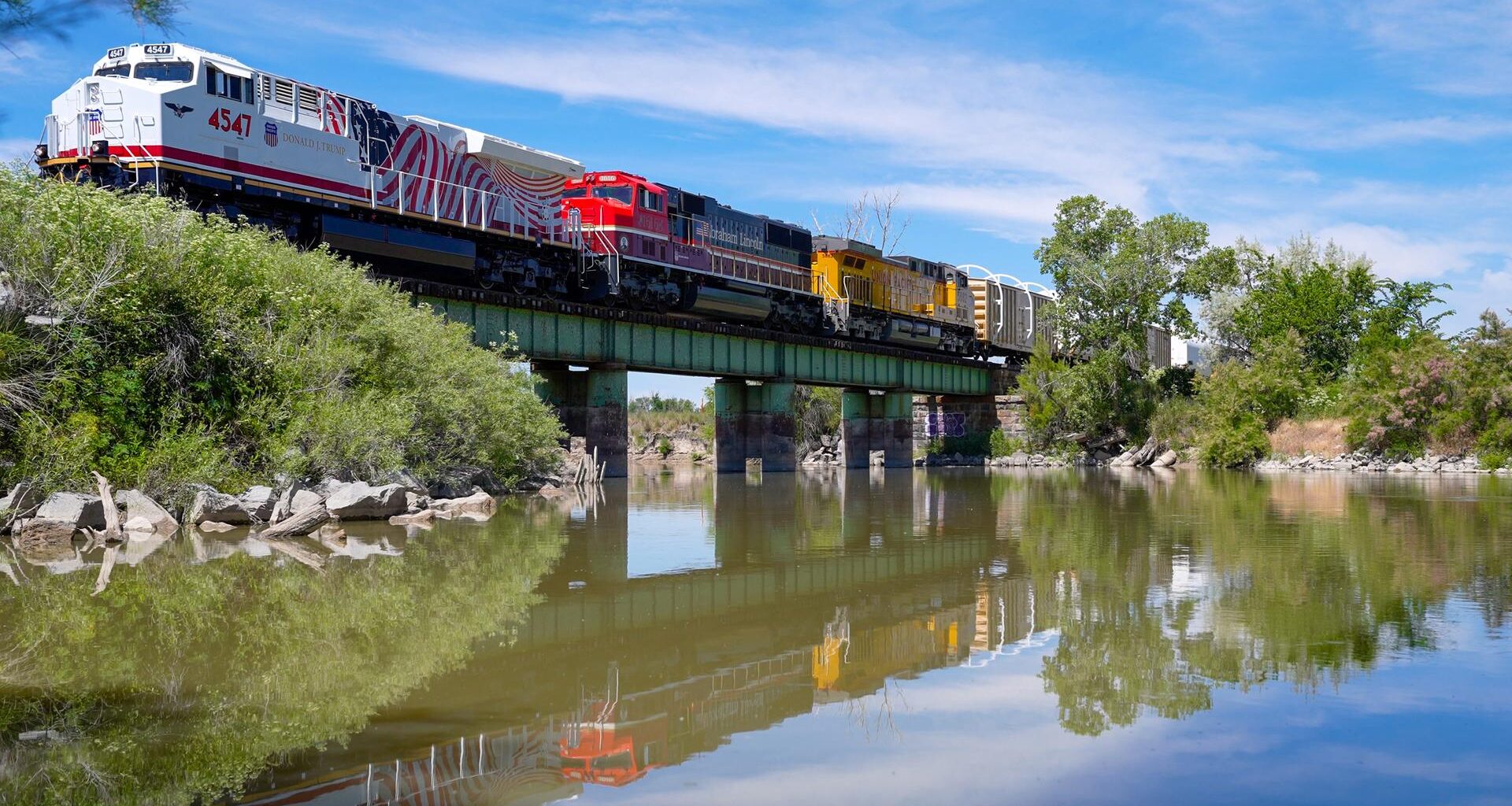 A train with many different colored cars crosses a green bridge over a body of water. There are trees along the shoreline.