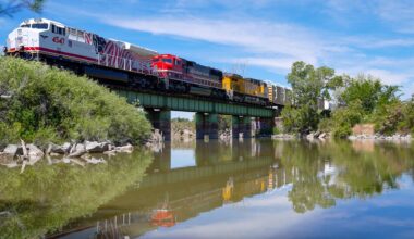 A train with many different colored cars crosses a green bridge over a body of water. There are trees along the shoreline.