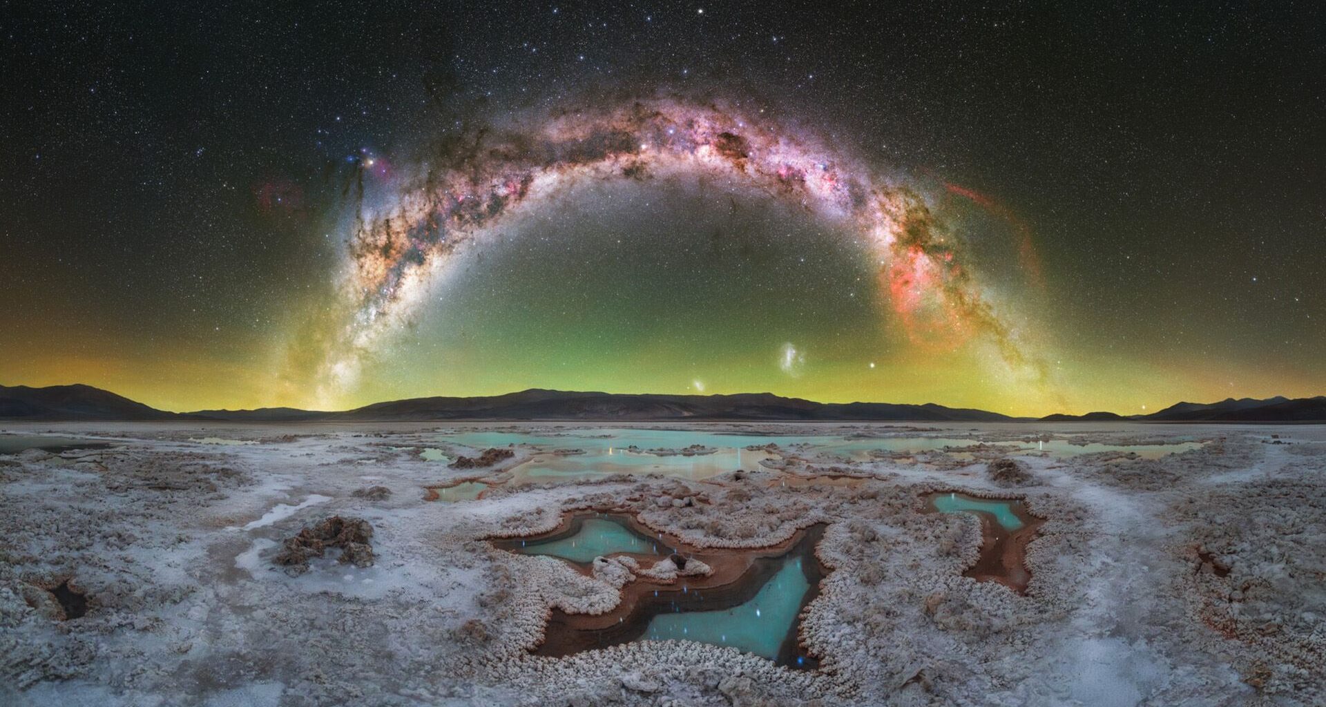 A panorama of the night sky showing the glowing arc of the Milky Way galaxy over a snowy landscape.