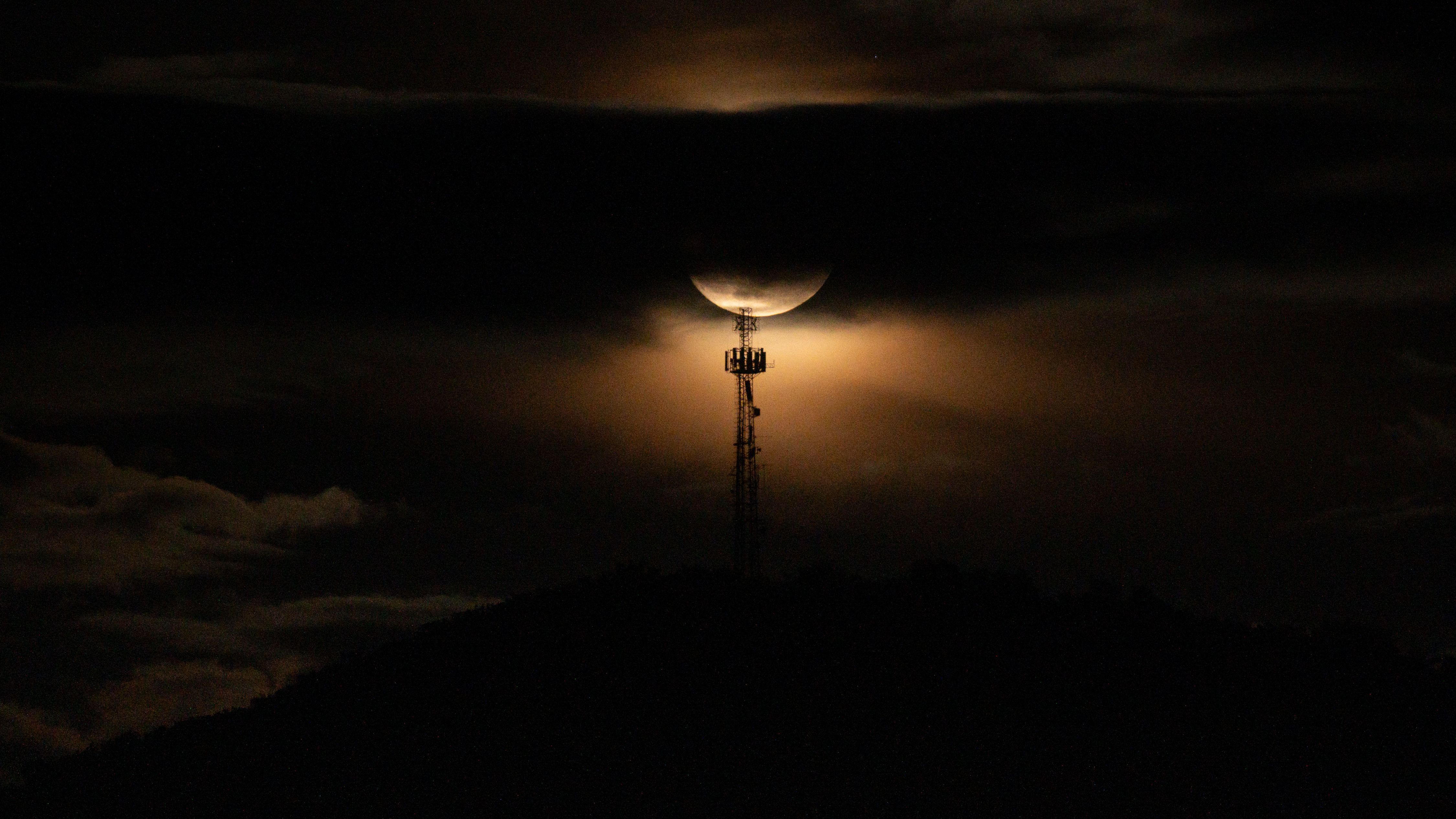 A yellow full moon is pictured in the night sky with its surface covered by a band of cloud. It's light shines out beneath to silhouette a communications tower.