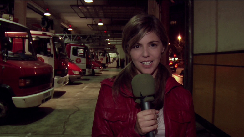 Manuela Velasco as Ángela Vidal in REC, standing in front of a line of ambulance cars and holding her microphone while she looks at the camera.