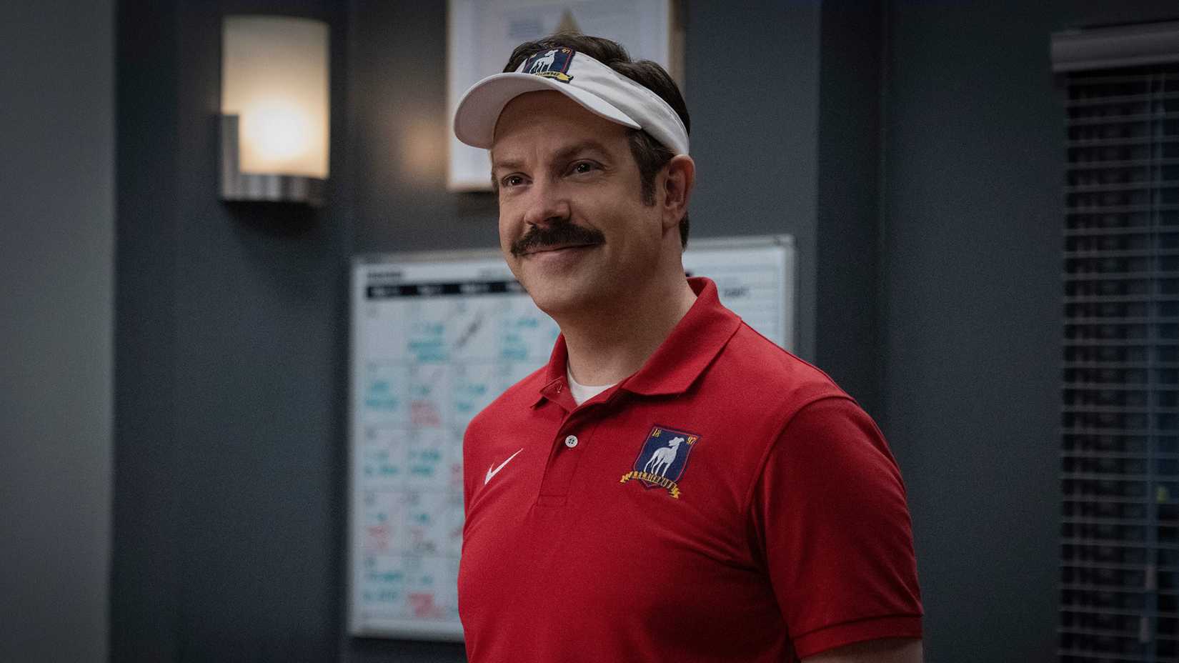 Ted standing in his office and smiling while wearing an A.F.C Richmond rimmed hat in Ted Lasso.