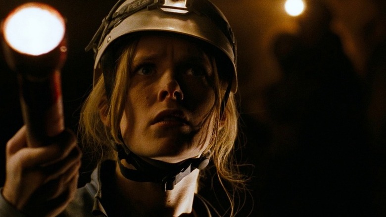 Shauna Macdonald as Sarah Carter in The Descent, wearing her spelunking helmet and holding out a flashlight while looking at something.