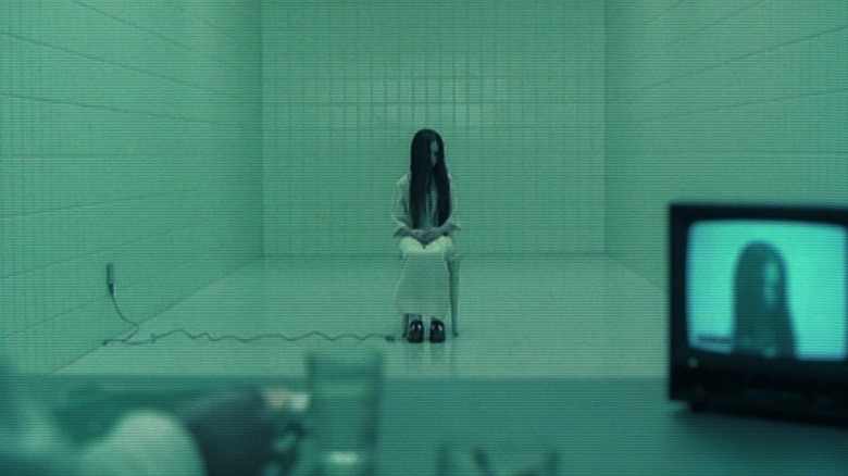 Daveigh Chase as Samara Morgan in The Ring, sitting in the center of a plain, tiled room, from the perspective of someone sitting at a desk with a small television broadcasting Samara's image on it.