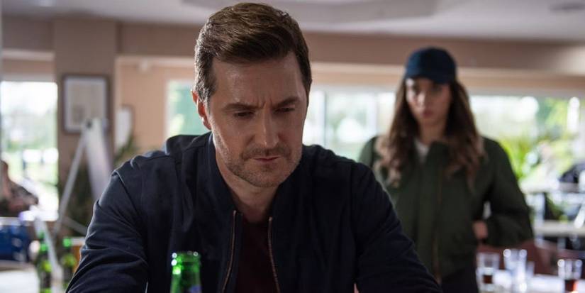 Richard Armitage sits at a counter with Hannah John-Kamen behind in The Stranger.