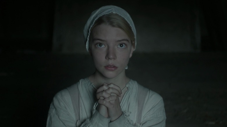 Anya Taylor-Joy as Thomasin in The Witch, kneeled down on the ground with her hands clasped in prayer as she looks on above her.