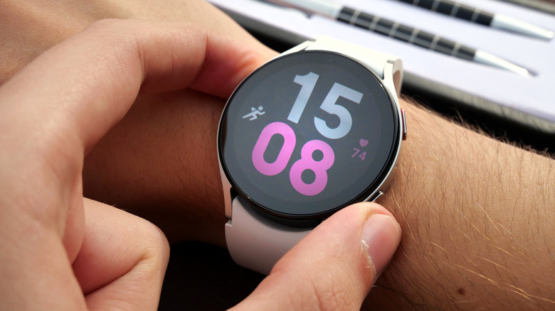 An individual using their left hand to adjust the Samsung smartwatch on their right wrist, the watch is displaying the time in a blue and pink font