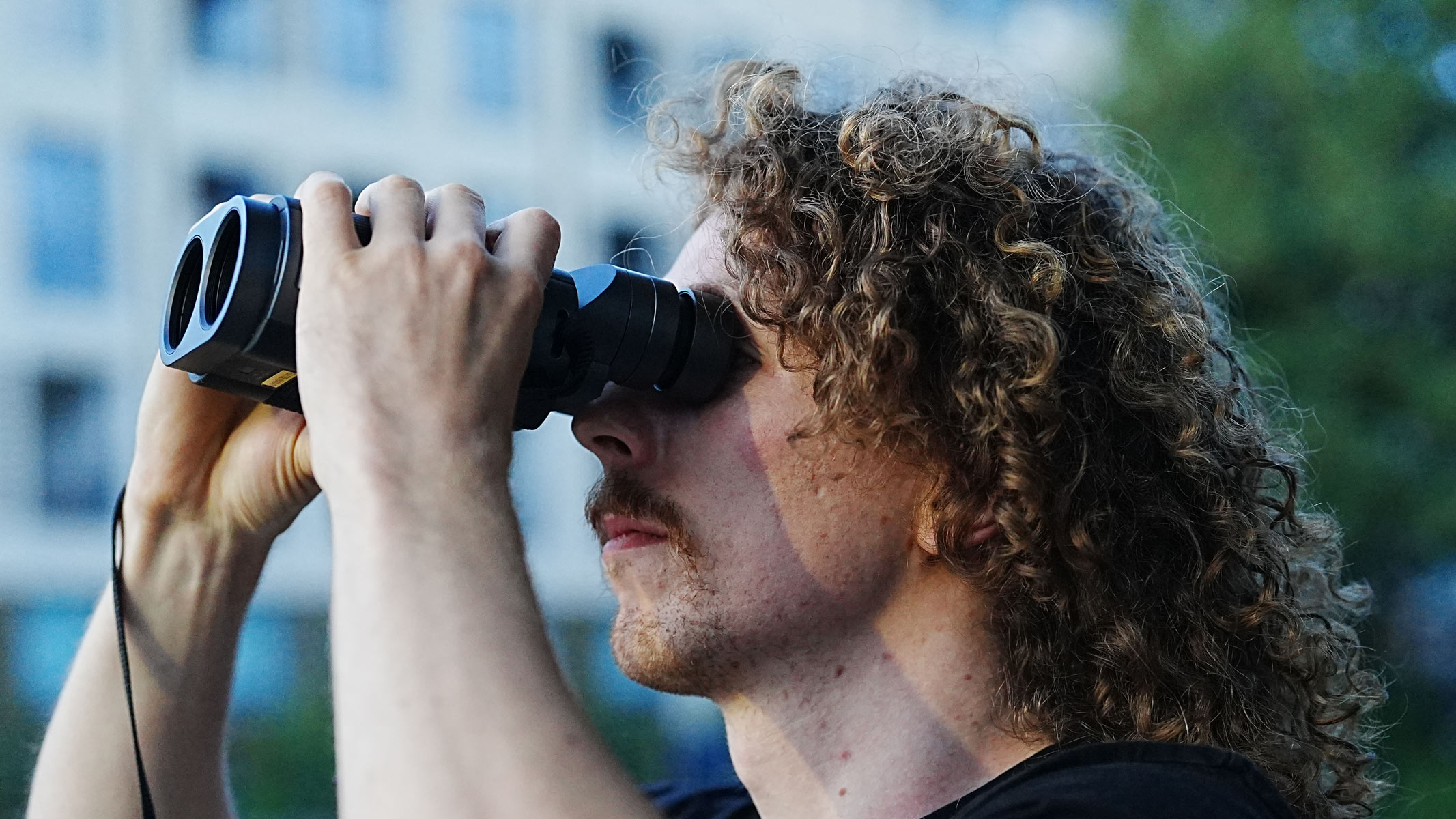 A close-up of a man looking through the Fujifilm Techno-Stabi TS-L 1640 image-stabilized binoculars with buildings and trees in the background.
