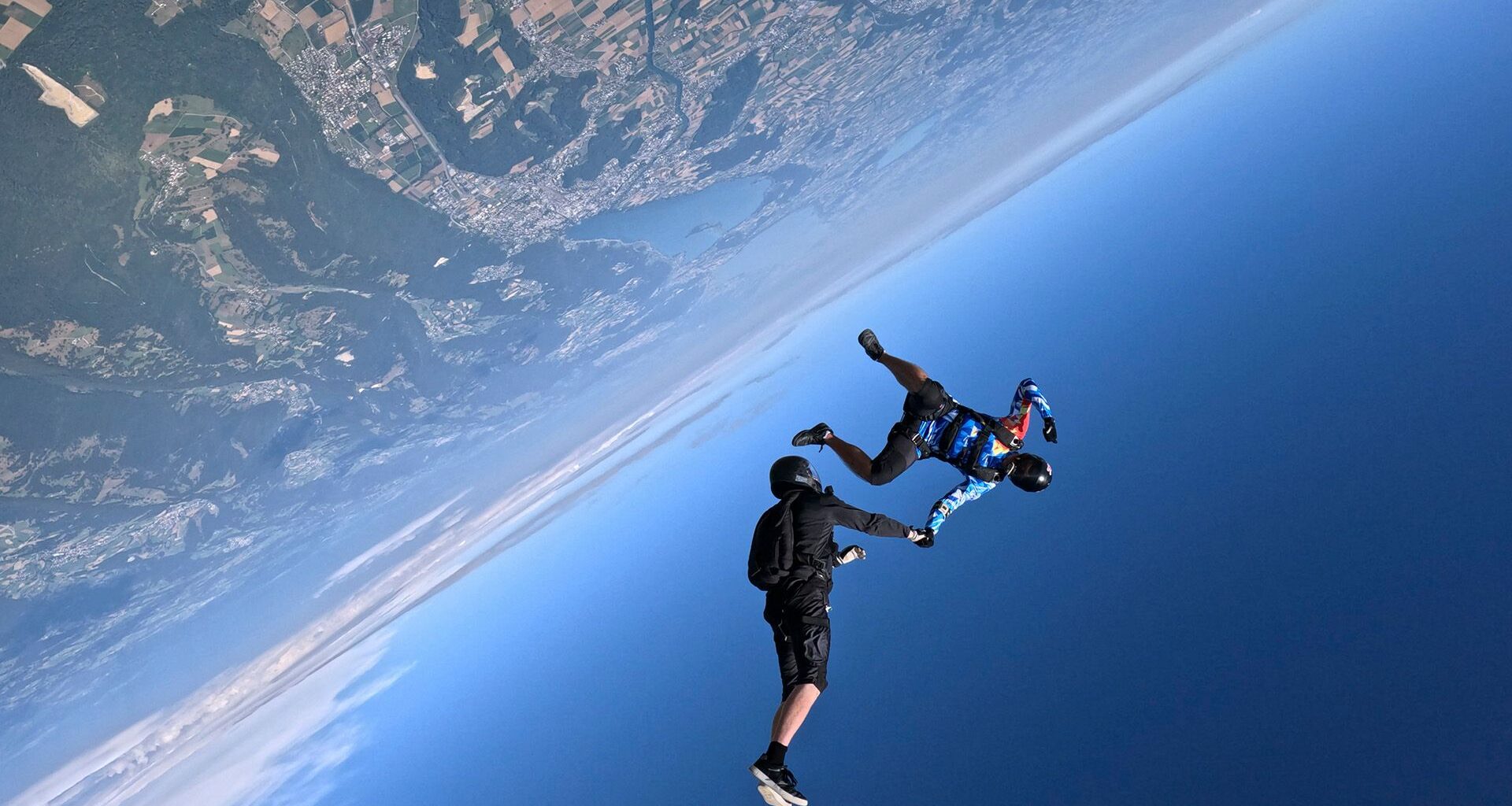 Two people hold hands while skydiving over Earth, a view of its surface next to the blue sky is seen.