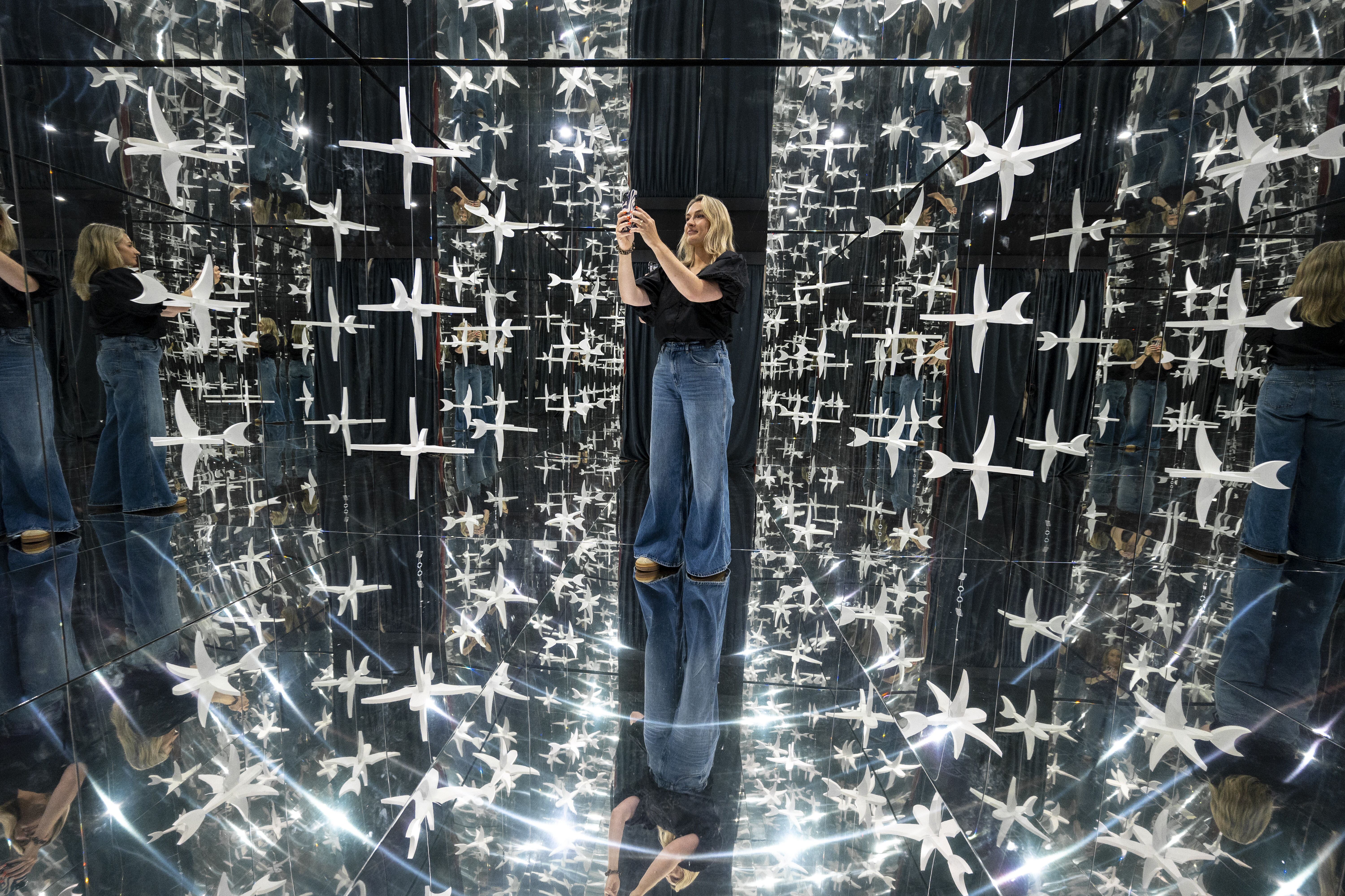 A visitor stands inside the exhibition's mirrored infinity room, surrounded by suspended white bird forms that multiply endlessly in every direction across the floor, walls and ceiling