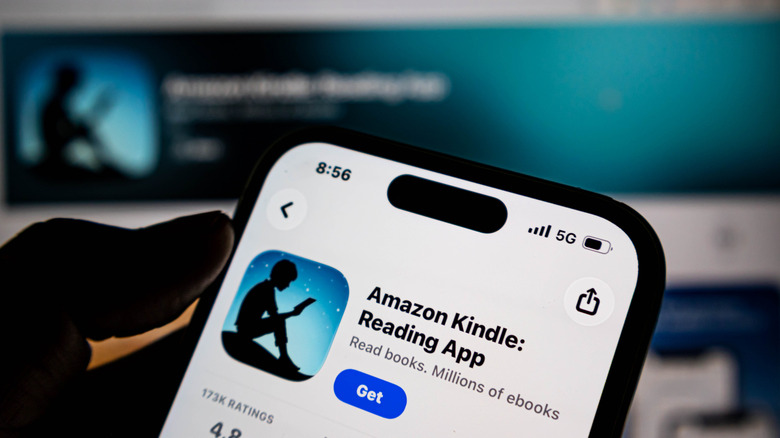 The silhouette of a hand holds a phone up with the Amazon Kindle app ready on their phone's pp store.